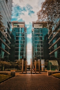 Two modern glass skyscrapers rise vertically, flanking a pathway with metallic pillars in the center. The reflections on the glass exteriors create a dynamic pattern under a partly cloudy sky. Trees are present on both sides, bringing a touch of greenery to the urban setting.