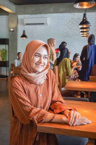 A woman wearing a tan hijab and a brown striped shirt is sitting at a wooden table in a modern cafe. She is smiling gently and there are several other people in the background, creating a lively atmosphere. The cafe interior features hanging pendant lights and a white brick wall.