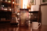 A cozy, warmly lit scene featuring a coffee setup. On the wooden table, there is a Chemex coffee maker containing brewed coffee next to a packaged coffee product with floral design and brand labeling. A small white espresso cup sits to the right, with a dimly visible kitchen background.