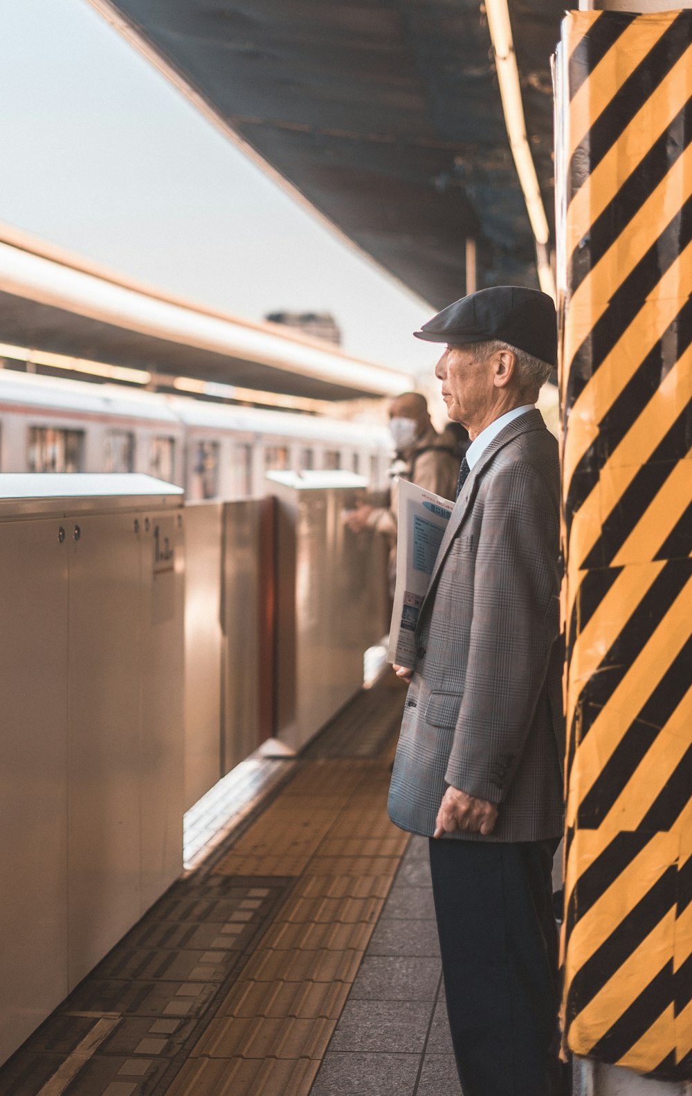 Mann in grauer Anzugjacke steht neben schwarz-gelb gestreiftem Pfosten am Bahnhof