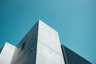 A minimalistic modern building with a concrete structure and a glass facade set against a clear blue sky. The architectural style is clean and geometric, emphasizing sharp lines and angles.