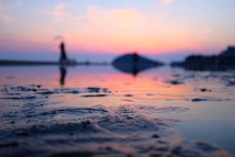 A calm and serene beach scene at sunset, with gentle waves reflecting the vibrant colors of the sky. The foreground is slightly blurred and the focus is on the texture of the wet sand. Silhouettes of people walking can be seen in the background, adding a sense of depth and tranquility to the image.