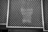 A metal grid with a honeycomb pattern covers a window, casting intricate shadows. The structure is framed by squared edges, and the overall appearance is monotonous due to the limited color range.