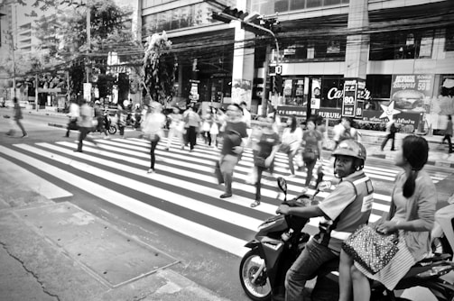 A busy urban scene featuring a group of people crossing a zebra-striped pedestrian crossing. The motion blur suggests movement and activity, while a man and woman are seated on a motorcycle waiting at the crosswalk. The background includes various storefronts and signage, indicating a commercial area.