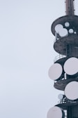 A tall structure featuring several large, circular satellite dishes attached to its sides, with a muted, overcast sky in the background. The structure appears industrial with metal railings and platforms.