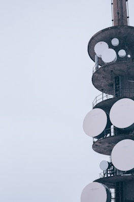 A tall structure featuring several large, circular satellite dishes attached to its sides, with a muted, overcast sky in the background. The structure appears industrial with metal railings and platforms.