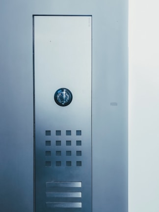 A metallic intercom panel is mounted on a wall, featuring a circular speaker at the top, a grid of small square openings below it, and two nameplates at the bottom.