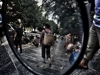 A bustling street market scene featuring people engaging in various activities, such as selling and buying goods. The street is lined with tall trees providing shade, and several individuals are carrying large woven baskets. Cars are parked along the pavement, and the atmosphere appears lively and dynamic.