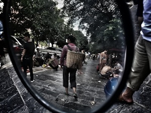 A bustling street market scene featuring people engaging in various activities, such as selling and buying goods. The street is lined with tall trees providing shade, and several individuals are carrying large woven baskets. Cars are parked along the pavement, and the atmosphere appears lively and dynamic.