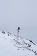 A snow-covered mountainous landscape with a metal pole topped by a small solar panel or device. A red and white windsock hangs from the pole, indicating wind direction. The surroundings are enveloped in fog, creating a serene and isolated atmosphere.