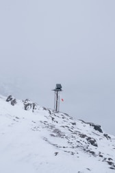 A snow-covered mountainous landscape with a metal pole topped by a small solar panel or device. A red and white windsock hangs from the pole, indicating wind direction. The surroundings are enveloped in fog, creating a serene and isolated atmosphere.