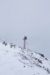 A snow-covered mountainous landscape with a metal pole topped by a small solar panel or device. A red and white windsock hangs from the pole, indicating wind direction. The surroundings are enveloped in fog, creating a serene and isolated atmosphere.