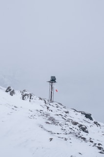 A snow-covered mountainous landscape with a metal pole topped by a small solar panel or device. A red and white windsock hangs from the pole, indicating wind direction. The surroundings are enveloped in fog, creating a serene and isolated atmosphere.