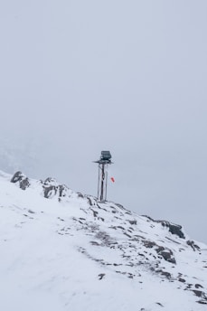 A snow-covered mountainous landscape with a metal pole topped by a small solar panel or device. A red and white windsock hangs from the pole, indicating wind direction. The surroundings are enveloped in fog, creating a serene and isolated atmosphere.