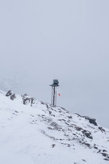 A snow-covered mountainous landscape with a metal pole topped by a small solar panel or device. A red and white windsock hangs from the pole, indicating wind direction. The surroundings are enveloped in fog, creating a serene and isolated atmosphere.
