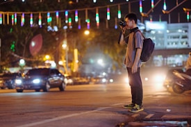 A person with a backpack is standing in the middle of a street at night, taking a photograph. The street is illuminated by various colorful lights and there are cars in motion nearby. The scene is vibrant and bustling with activity.