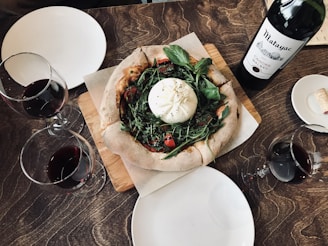 A rustic table setting featuring a fresh pizza topped with arugula, tomatoes, and a large ball of burrata cheese. Surrounding the pizza are several glasses of red wine and a bottle of wine with a label visible. The table setting includes minimal white plates and a cork beside the bottle.