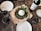 A rustic table setting featuring a fresh pizza topped with arugula, tomatoes, and a large ball of burrata cheese. Surrounding the pizza are several glasses of red wine and a bottle of wine with a label visible. The table setting includes minimal white plates and a cork beside the bottle.