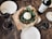 A rustic table setting featuring a fresh pizza topped with arugula, tomatoes, and a large ball of burrata cheese. Surrounding the pizza are several glasses of red wine and a bottle of wine with a label visible. The table setting includes minimal white plates and a cork beside the bottle.