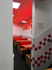 A diner with red and white checkered tile walls, red cushioned booth seating, and a sign with text. The lighting casts a warm red glow across the ceiling and walls.