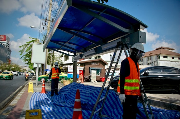 Construction workers wearing protective gear and high-visibility vests are working at a bus stop under a clear blue sky. Traffic cones and a blue tarpaulin cover the ground, and a ladder is set up next to a new shelter structure. Vehicles are visible on the busy street in the background.