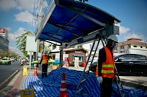 Construction workers wearing protective gear and high-visibility vests are working at a bus stop under a clear blue sky. Traffic cones and a blue tarpaulin cover the ground, and a ladder is set up next to a new shelter structure. Vehicles are visible on the busy street in the background.