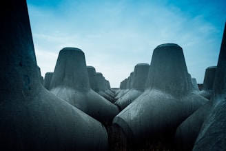 Massive, grey concrete tetrapods are arranged closely together, stretching towards the horizon under a sky that is partly overcast with patches of blue. The structures appear imposing and function as wave breakers, typically used in coastal engineering.