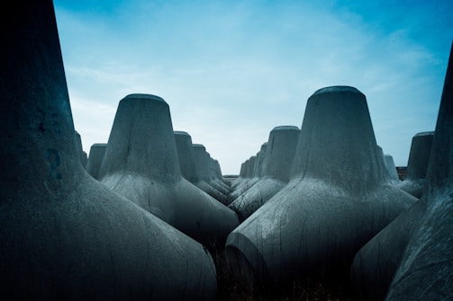 Massive, grey concrete tetrapods are arranged closely together, stretching towards the horizon under a sky that is partly overcast with patches of blue. The structures appear imposing and function as wave breakers, typically used in coastal engineering.