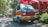 A colorful van with a retro design sits parked on a sunny street surrounded by lush greenery. The vehicle is painted in shades of red, yellow, and blue, contrasting with its natural surroundings. People are seen walking in the background, near market stalls or park pathways under a canopy of vibrant red structures.