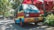 A colorful van with a retro design sits parked on a sunny street surrounded by lush greenery. The vehicle is painted in shades of red, yellow, and blue, contrasting with its natural surroundings. People are seen walking in the background, near market stalls or park pathways under a canopy of vibrant red structures.