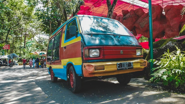 A colorful van with a retro design sits parked on a sunny street surrounded by lush greenery. The vehicle is painted in shades of red, yellow, and blue, contrasting with its natural surroundings. People are seen walking in the background, near market stalls or park pathways under a canopy of vibrant red structures.