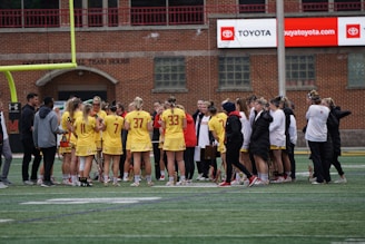 A group of athletes in yellow jerseys and shorts gather on a sports field, surrounded by coaches and team staff dressed in winter clothing. The setting appears to be a team huddle during a sports event, with a goalpost in the background and an advertisement for Toyota on a brick building nearby.