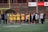 A group of athletes in yellow jerseys and shorts gather on a sports field, surrounded by coaches and team staff dressed in winter clothing. The setting appears to be a team huddle during a sports event, with a goalpost in the background and an advertisement for Toyota on a brick building nearby.