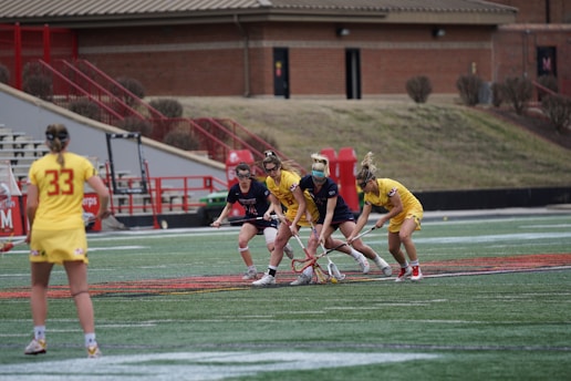 A group of female lacrosse players is engaged in play on a sports field. Two teams are involved, one wearing yellow uniforms and the other wearing dark blue. The players are focused on the ball on the ground, using their lacrosse sticks to gain possession. The background shows a stadium with tiered seating and a brick building.