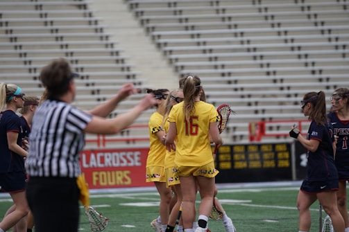 A group of female lacrosse players are gathered on a sports field. The players are wearing protective goggles and uniforms; one team is in yellow and the other is in dark blue. A referee is in the foreground wearing a black and white striped shirt. The stadium's seats are mostly empty, and there are signs and banners in the background.