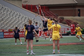A women's lacrosse match is in progress on a sports field. Two players from opposing teams are jumping mid-air, reaching for the ball with their lacrosse sticks, while a referee observes closely. The teams are dressed in contrasting uniforms, one in red and navy, and the other in yellow. Several other players are visible on the field, and empty bleachers are seen in the background.