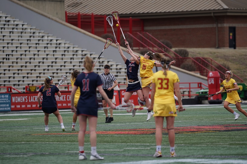 A women's lacrosse match is in progress on a sports field. Two players from opposing teams are jumping mid-air, reaching for the ball with their lacrosse sticks, while a referee observes closely. The teams are dressed in contrasting uniforms, one in red and navy, and the other in yellow. Several other players are visible on the field, and empty bleachers are seen in the background.