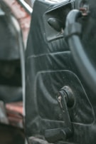 Interior detail of an older car door with a manual window roller and worn black upholstery. The focus is on the door panel, showing signs of age and use.