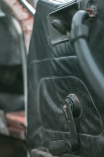 Interior detail of an older car door with a manual window roller and worn black upholstery. The focus is on the door panel, showing signs of age and use.