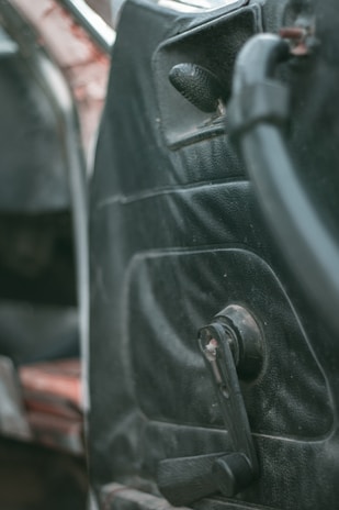 Interior detail of an older car door with a manual window roller and worn black upholstery. The focus is on the door panel, showing signs of age and use.