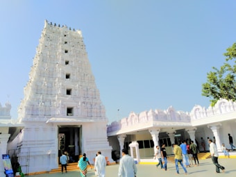 A large white temple tower with intricate carvings and statues rises against a clear blue sky. People, both men and women, are walking around the area, some entering the temple. A tree is visible on the right side, adding greenery to the scene.