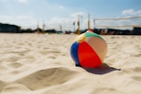 A colorful beach ball lies on sandy ground with beach volleyball nets in the background, under a clear blue sky with a few clouds.