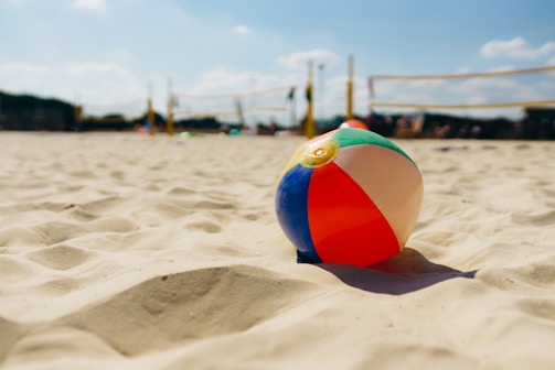 A colorful beach ball lies on sandy ground with beach volleyball nets in the background, under a clear blue sky with a few clouds.