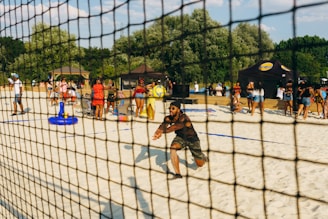 A man is playing beach volleyball, crouched in an action pose, surrounded by a busy scene with numerous people enjoying a sunny day on the sandy court. There are tents in the background and groups of people are standing, sitting on chairs, and interacting. The setting is festive and lively, with green trees framing the area and a clear blue sky overhead.