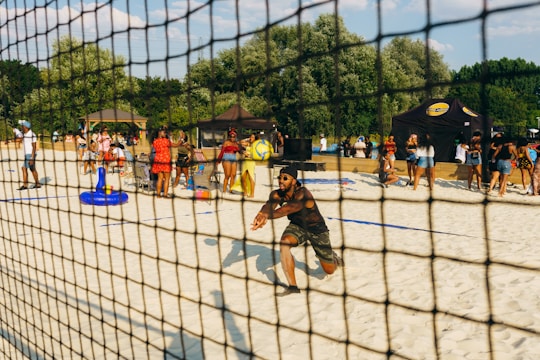 A man is playing beach volleyball, crouched in an action pose, surrounded by a busy scene with numerous people enjoying a sunny day on the sandy court. There are tents in the background and groups of people are standing, sitting on chairs, and interacting. The setting is festive and lively, with green trees framing the area and a clear blue sky overhead.