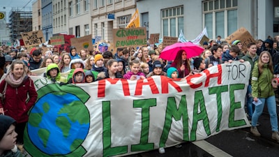 A large group of people, mostly young individuals, are participating in a climate march. They hold banners and signs with slogans advocating for environmental action. One prominent banner reads 'March for Climate' with a drawing of the Earth. Some participants are wearing colorful clothing, and one person holds a bright pink umbrella.
