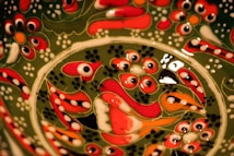 Intricate, colorful ceramic design featuring floral and abstract patterns with vibrant red, green, white, and black accents. The motifs are densely packed, creating a rich and ornate visual texture.