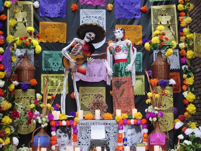A colorful Day of the Dead altar features two decorated skeleton figures, one playing a guitar. Vibrant papel picado with intricate patterns hang in the background. The altar is adorned with marigolds, candles, and photographs, surrounded by baskets and colorful flowers.