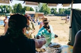 People are gathered at an outdoor event, possibly a market or festival. A person in a turquoise outfit is reaching for food from a vendor's stall, while another person nearby is holding a bottled water. There are several tables and people in the background enjoying the sunny day, with trees and a building in the distance.