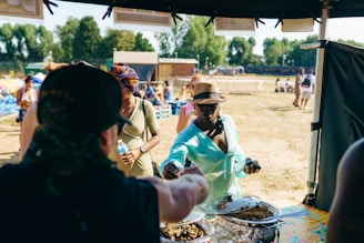 People are gathered at an outdoor event, possibly a market or festival. A person in a turquoise outfit is reaching for food from a vendor's stall, while another person nearby is holding a bottled water. There are several tables and people in the background enjoying the sunny day, with trees and a building in the distance.